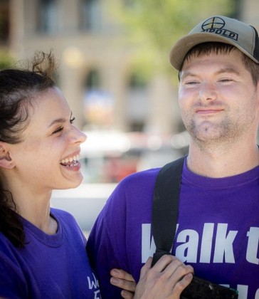 Two people wearing purple shirts that read 'Walk to...' smiling and sharing a moment together. A woman with dark hair is laughing while a man wearing a baseball cap looks content.